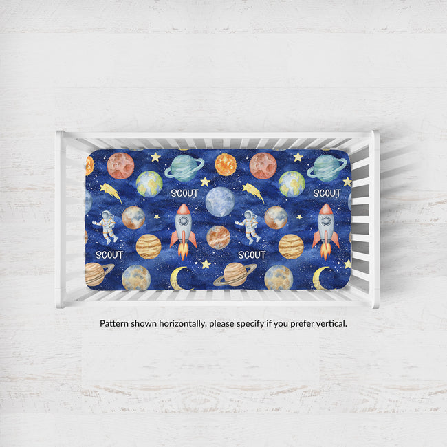 Overhead view of a white crib with the Space Explorers personalized sheet, showing the navy space pattern and Scout lettering oriented horizontally.
