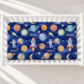 A fitted crib sheet with a colorful space-themed print featuring planets, stars, astronauts, and rockets on a dark blue background, shown inside a white wooden crib.