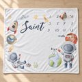 A square white baby blanket with a space-themed design, featuring watercolor illustrations of astronauts, planets, stars, and rockets, with a customizable 'Saint [name] months' text printed on the front. The blanket is laid flat on a light wood floor.