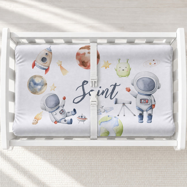 A white personalized changing pad cover with a space-themed design, featuring watercolor astronauts, planets, stars, and the name 'Saint' in script. 