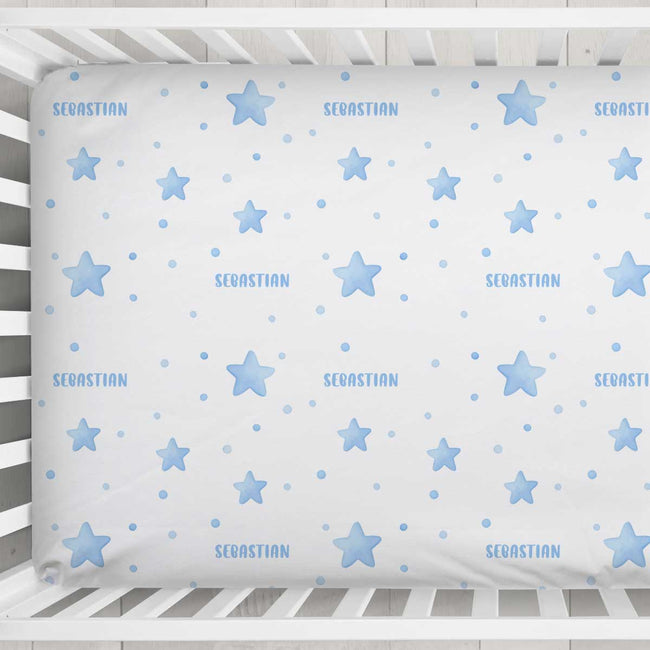 A white fitted crib sheet with a light blue pattern of stars, dots, and the name 'SEBASTIAN' printed repeatedly, shown inside a white wooden crib.