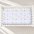 A white crib mattress fitted with a white sheet printed with light blue stars, dots, and the name 'SEBASTIAN' repeated across the surface, shown inside a white crib on a light neutral floor.