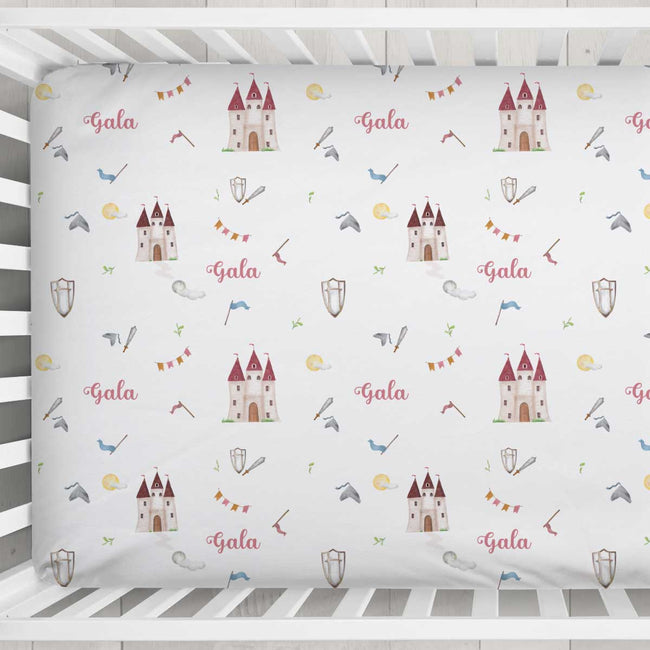A fitted crib sheet with a white background and a repeating pattern of fairytale castles, bows, arrows, flags, and the word 'Gala' in pink script, shown inside a white wooden crib.
