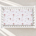 A fitted crib mattress with a white background and a repeating galah theme, including castles, bows, arrows, shields, and the word 'Gala' in red script, shown inside a white wooden crib.