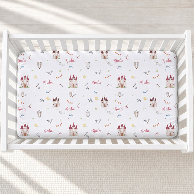 A fitted crib mattress with a white background and a repeating galah theme, including castles, bows, arrows, shields, and the word 'Gala' in red script, shown inside a white wooden crib.