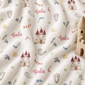 A close-up of a white fabric with a playful medieval theme print, featuring watercolor-style castles, swords, shields, flags, and the word 'Gala' in pink script. A small wooden toy castle is placed on the right side of the frame.