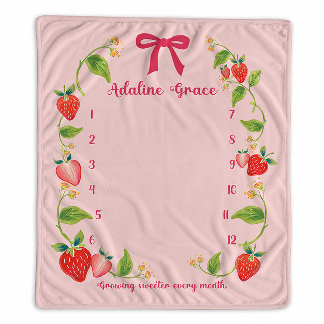 A personalized baby milestone blanket featuring a pink background decorated with a strawberry and green leaf border, a pink bow at the top, and the text 'Adaline Grace' along with a numbered countdown from 1 to 12.