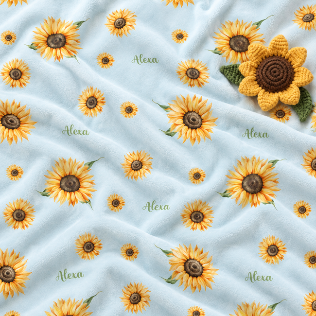 A light blue plush blanket with a repeating sunflower pattern, featuring the name 'Alexa' printed in green script between the flowers. A crocheted yellow sunflower is placed on the top right corner of the blanket.
