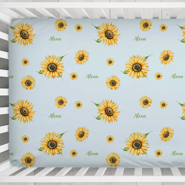 A personalized baby crib mattress cover featuring a light blue background with a repeating pattern of yellow sunflowers and the name 'Alexa' printed in green script. The cover is shown fitted inside a white wooden crib.