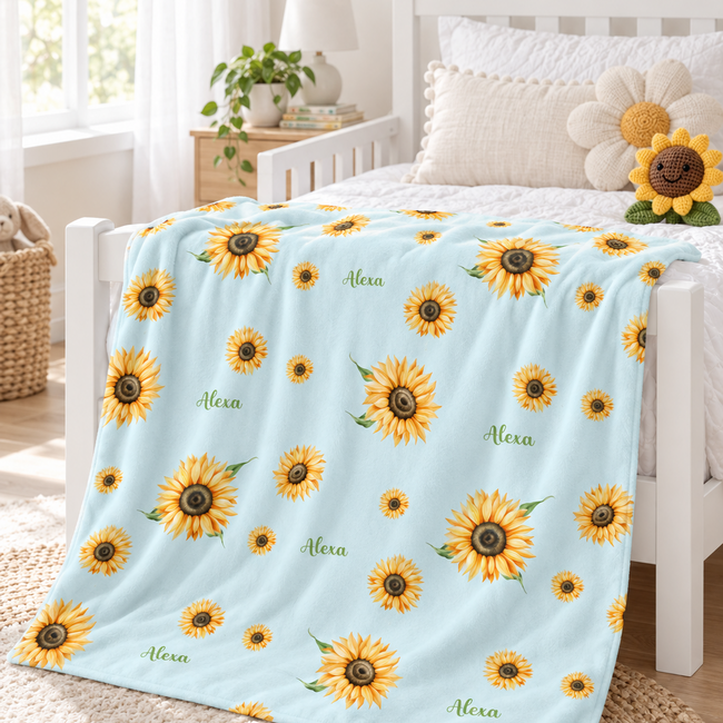 A light blue blanket with a yellow sunflower pattern and the name 'Alexa' printed in green script, draped over a white bed in a bright, sunlit bedroom.
