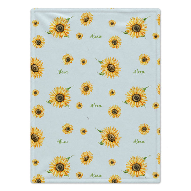 A square fabric item, likely a small blanket or towel, featuring a repeating pattern of yellow sunflowers and the name 'Alexa' in green cursive on a light blue background.