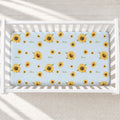 A light blue crib mattress cover featuring a repeating pattern of yellow sunflowers and the name 'Alexa' in green script, shown inside a white wooden baby crib.