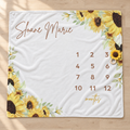 A personalized white blanket or mat laid flat on a light wood floor, printed with yellow sunflower illustrations, the name 'Sloane Marie' in brown script, a 12-month calendar grid, and 'months' in gold script.