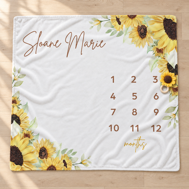 A personalized white blanket or mat laid flat on a light wood floor, printed with yellow sunflower illustrations, the name 'Sloane Marie' in brown script, a 12-month calendar grid, and 'months' in gold script.