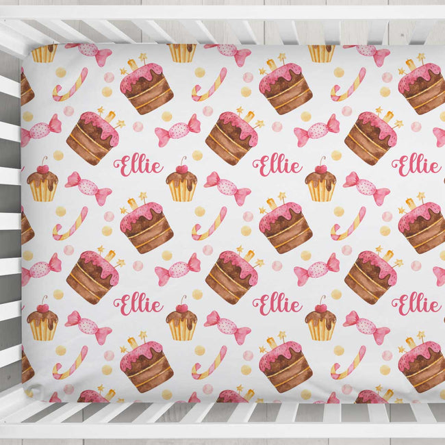 A crib mattress fitted sheet with a repeating pattern of watercolor cupcakes, pink lollipops, candy canes, and the name 'Ellie' in pink script, shown on a white wooden crib.