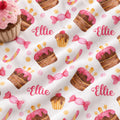 A close-up of a white fabric printed with a repeating pattern of pink-frosted cupcakes, pink candies, yellow dots, and the name 'Ellie' in pink script. A plush pink cupcake prop is visible in the top-left corner.