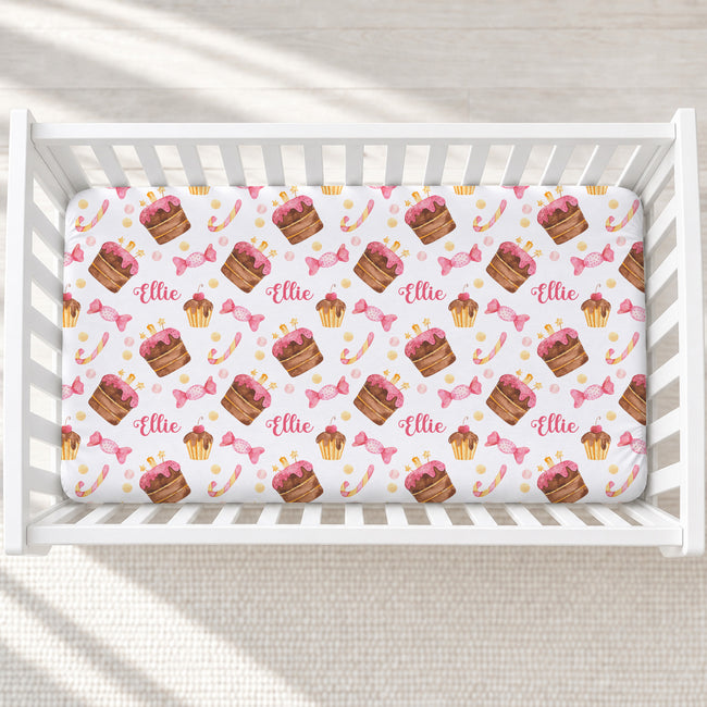 A fitted crib sheet with a repeating pattern of cupcakes, pink bows, candy canes, and the name 'Ellie' in script, shown inside a white wooden crib on a light floor.