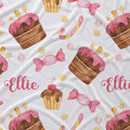 A white fabric blanket or throw featuring a whimsical watercolor pattern of pink-frosted cupcakes, candy canes, pink wrapped sweets, gold stars, and the name 'Ellie' in pink script.