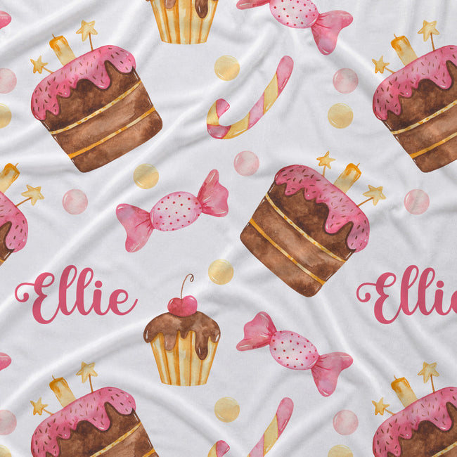A white fabric blanket or throw featuring a whimsical watercolor pattern of pink-frosted cupcakes, candy canes, pink wrapped sweets, gold stars, and the name 'Ellie' in pink script.