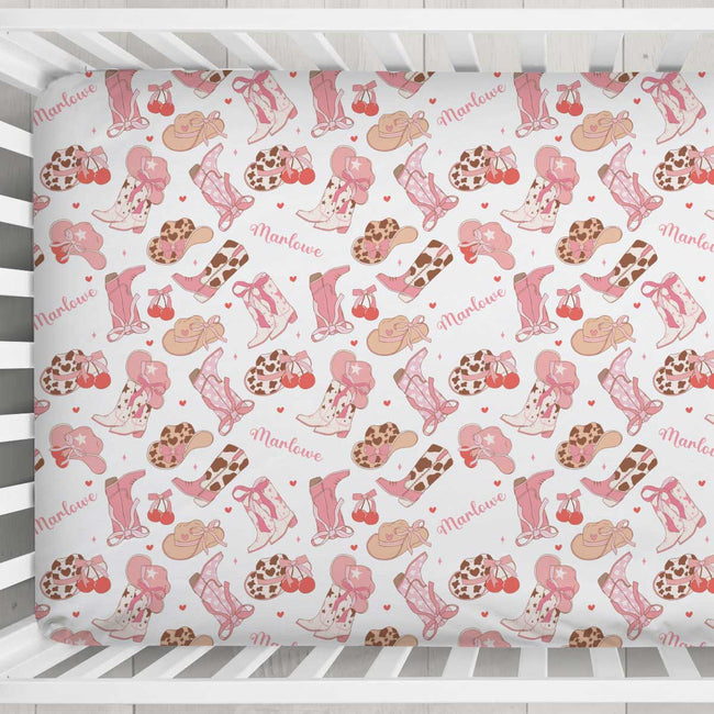 A fitted crib sheet with a western-themed pattern featuring pink cowboy boots, tan hats, brown leopard-print boots, cherries, and the word 'Marvelue' in script, shown inside a white wooden crib.