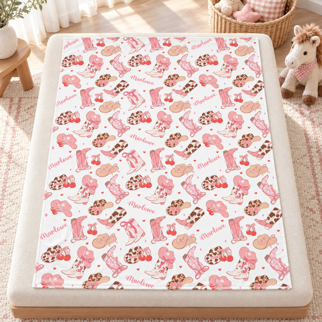 Personalized Cowgirl Boots Baby Girl Blanket. Pink cowgirl boots, western hats, bows, cherries, and hearts surround a custom name on a white background.