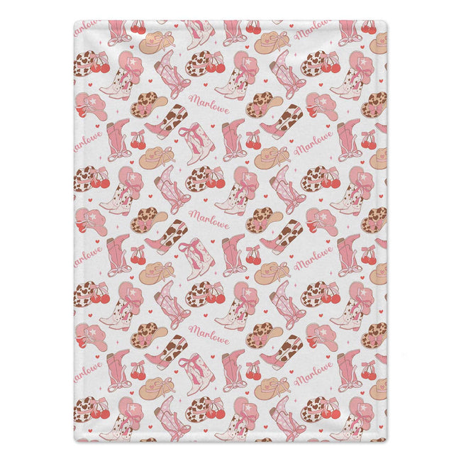 Personalized Cowgirl Boots Baby Girl Blanket. Pink cowgirl boots, western hats, bows, cherries, and hearts surround a custom name on a white background.