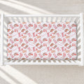 A fitted crib mattress with a white background and a pink and brown whimsical pattern of boots, hats, and the word 'Marlowe' printed repeatedly, shown inside a white wooden crib.