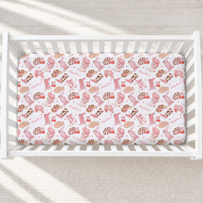 A fitted crib mattress with a white background and a pink and brown whimsical pattern of boots, hats, and the word 'Marlowe' printed repeatedly, shown inside a white wooden crib.