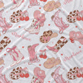 A close-up of a white fabric with a repeating western-themed pattern featuring pink cowboy boots, tan cowboy hats with hearts, brown-and-white cow-print hats, red cherries, small pink hearts, and the word 'Marlowe' in pink script.