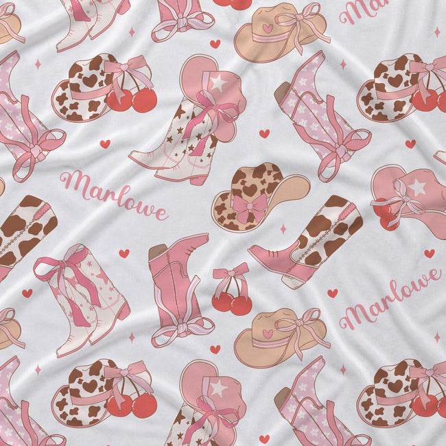 A close-up of a white fabric with a repeating western-themed pattern featuring pink cowboy boots, tan cowboy hats with hearts, brown-and-white cow-print hats, red cherries, small pink hearts, and the word 'Marlowe' in pink script.