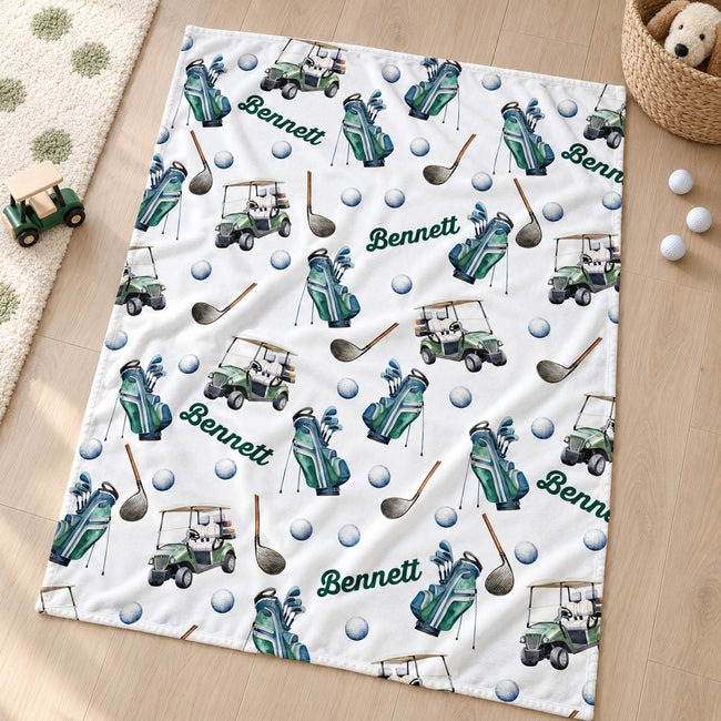 A personalized golf-themed blanket laid out on a light wood floor, featuring a repeating pattern of green and blue golf bags, golf carts, golf balls, and mallets, with the name 'Bennett' printed in green. A small toy golf cart and golf balls are placed nearby as props.