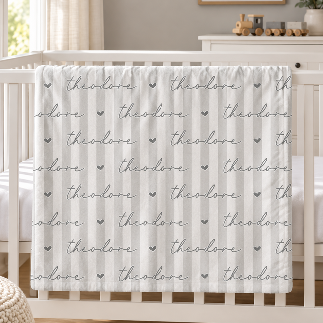 A white crib bedding set with vertical stripes and the name 'theodore' printed repeatedly in gray script, styled in a bright nursery setting.