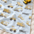 A personalized baby blanket with a light blue background and a yellow construction vehicle pattern, featuring the name 'DANIEL' printed in black. A small yellow toy dump truck is placed on top of the blanket, which is laid out on a beige fabric surface.