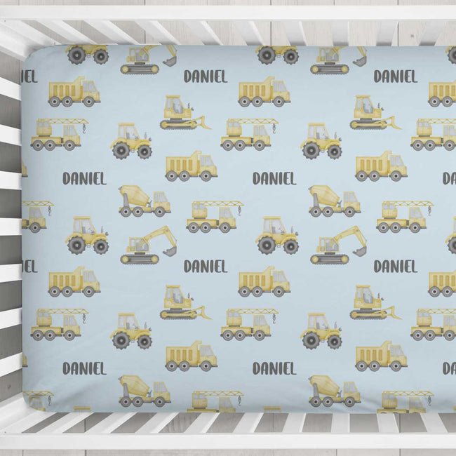 A personalized baby crib mattress cover with a light blue background, featuring a repeating pattern of yellow construction vehicles and the name 'DANIEL' in dark gray. The cover is shown fitted inside a white wooden crib.