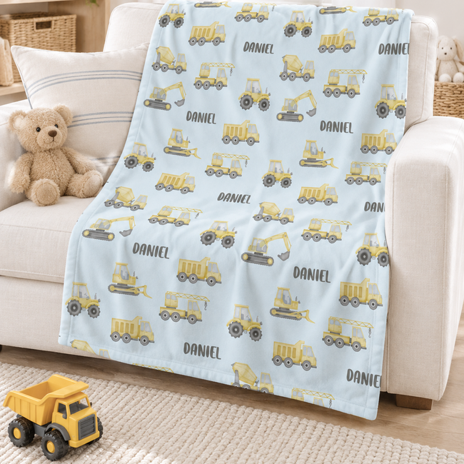 A personalized baby blanket with a light blue background and a yellow construction vehicle pattern, printed with the name 'DANIEL', draped over a white armchair in a bright nursery setting.