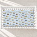 A baby crib with white slats, fitted with a light blue sheet printed with yellow construction vehicles and the name 'DANIEL' repeated across the surface.