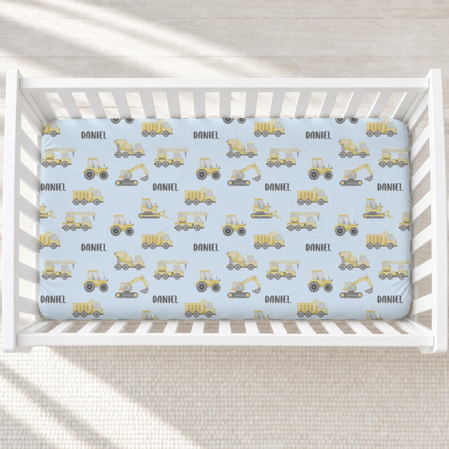 A baby crib with white slats, fitted with a light blue sheet printed with yellow construction vehicles and the name 'DANIEL' repeated across the surface.