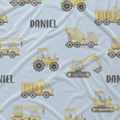 Light blue fabric with a repeating pattern of yellow construction vehicles including tractors, dump trucks, excavators, and cranes, with the name 'DANIEL' printed in gray.