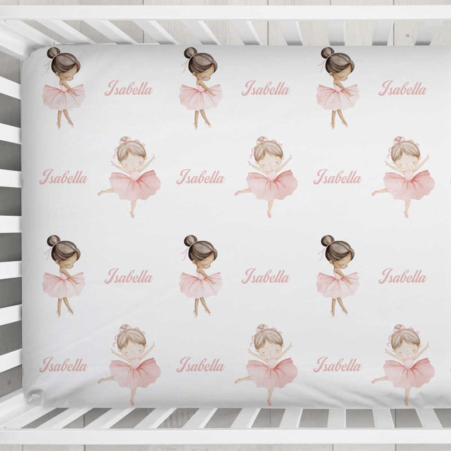 A baby crib sheet featuring a repeating pattern of ballerina illustrations and the name 'Isabella' in pink script, shown inside a white wooden crib.