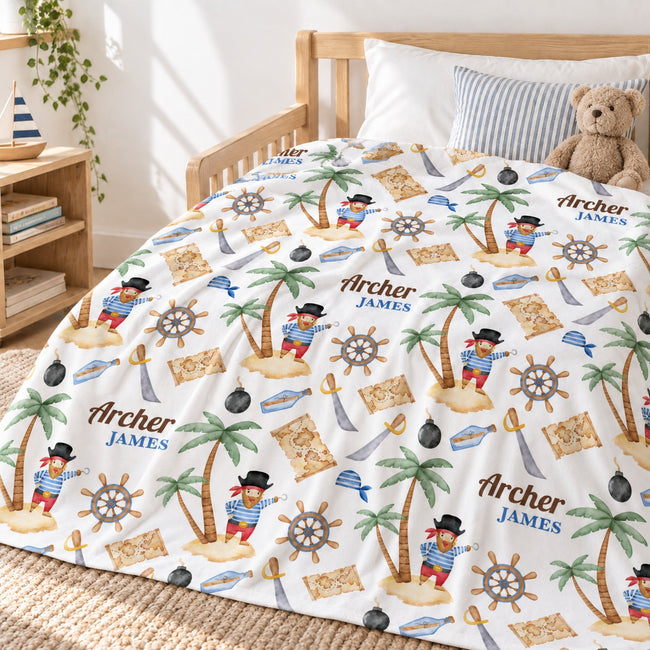 A personalized blanket featuring a pirate-themed pattern of palm trees, treasure maps, ship's wheels, swords, and characters in red and blue outfits. The duvet cover is personalized with the name 'Archer JAMES' in brown text.