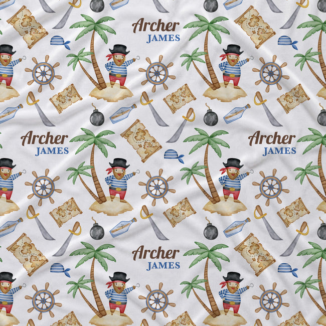 A close-up of a white fabric surface printed with a repeating pirate-themed pattern. The design includes palm trees on small islands, treasure maps, ship's wheels, swords, bottles, and a recurring figure labeled 'Archer JAMES'.