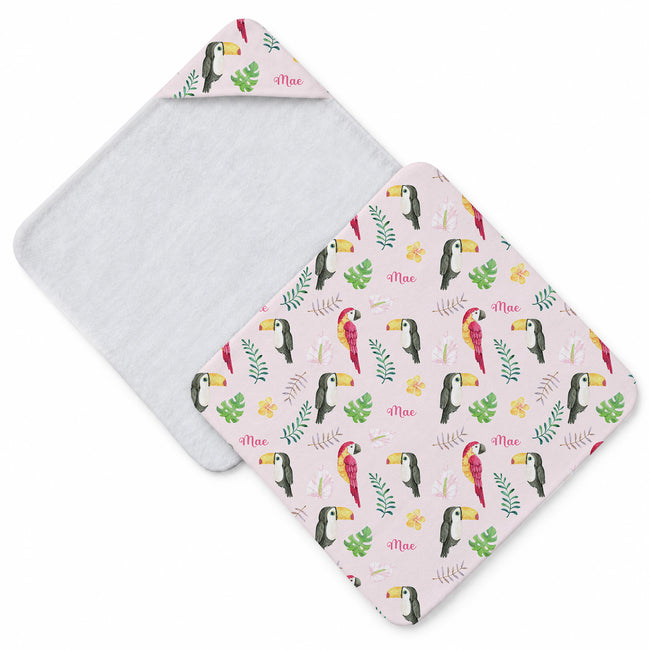 A baby hooded towel shown flat on a white background, with one half folded over to reveal a white plush lining and the other half showing a pale pink printed exterior with toucan and tropical leaf motifs and the name 'Mae'.