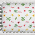 A fitted sheet with a tropical floral pattern featuring pink plumeria, yellow and white plumeria, green palm leaves, and the text 'Daisy MAE' in red and green, shown inside a white wooden crib.