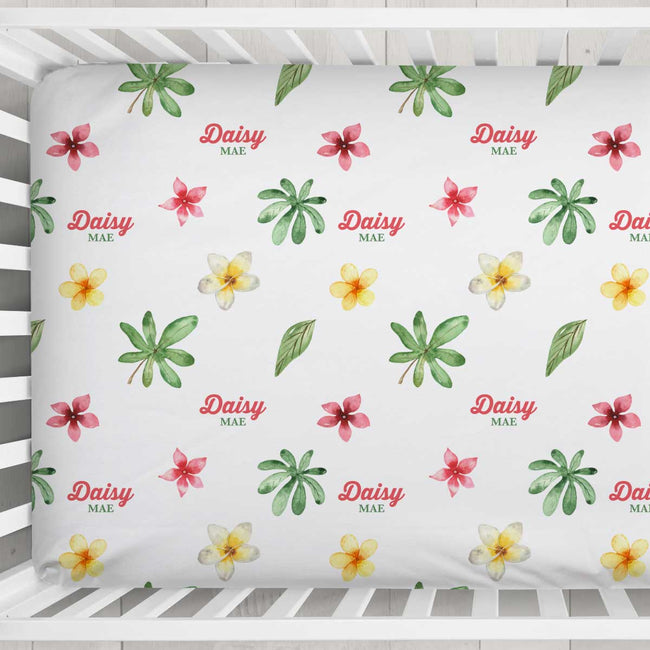 A fitted sheet with a tropical floral pattern featuring pink plumeria, yellow and white plumeria, green palm leaves, and the text 'Daisy MAE' in red and green, shown inside a white wooden crib.