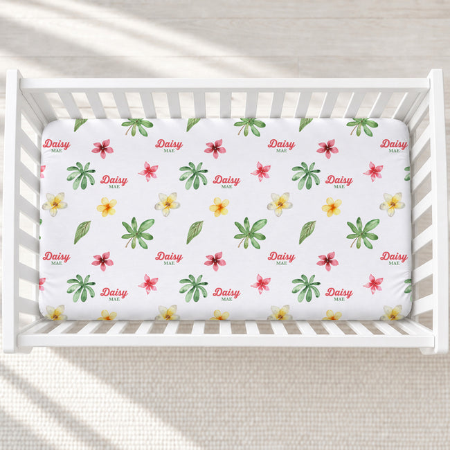 A white crib mattress fitted sheet with a colorful floral and leaf pattern, featuring the word 'Daisy' repeated in red script. The crib is shown from a top-down view on a light beige textured blanket.