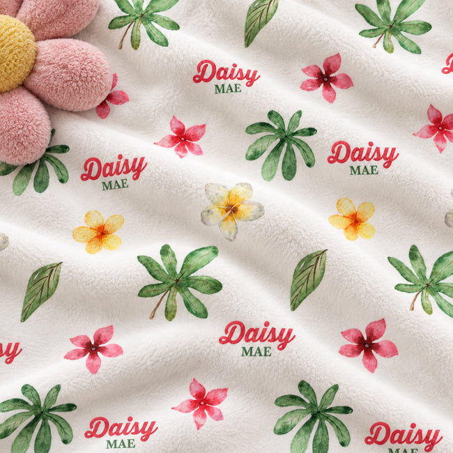A close-up of a white fabric with a repeating tropical print featuring green palm leaves, pink hibiscus flowers, yellow flowers, and the text 'Daisy MAE' in red and green. A plush pink flower toy is partially visible in the top-left corner.