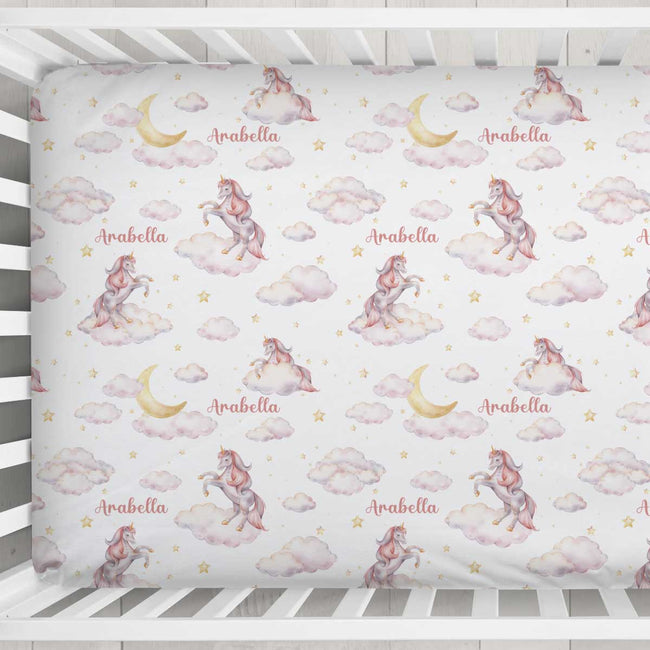 A crib mattress or fitted sheet with a repeating whimsical print of pink and white unicorns on clouds, yellow crescent moons, small gold stars, and the name 'Arabella' in script. The bedding is shown inside a white wooden crib.