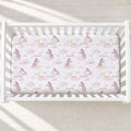 A fitted crib mattress with a whimsical fairy and cloud print, shown inside a white wooden crib on a neutral textured floor.