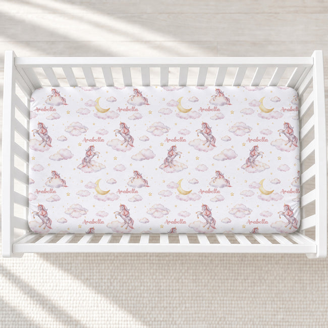 A fitted crib mattress with a whimsical fairy and cloud print, shown inside a white wooden crib on a neutral textured floor.
