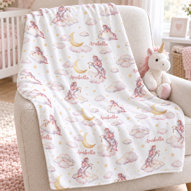 A white baby blanket with a pink and gold unicorn and moon pattern is draped over a beige armchair in a nursery setting, next to a plush unicorn toy.